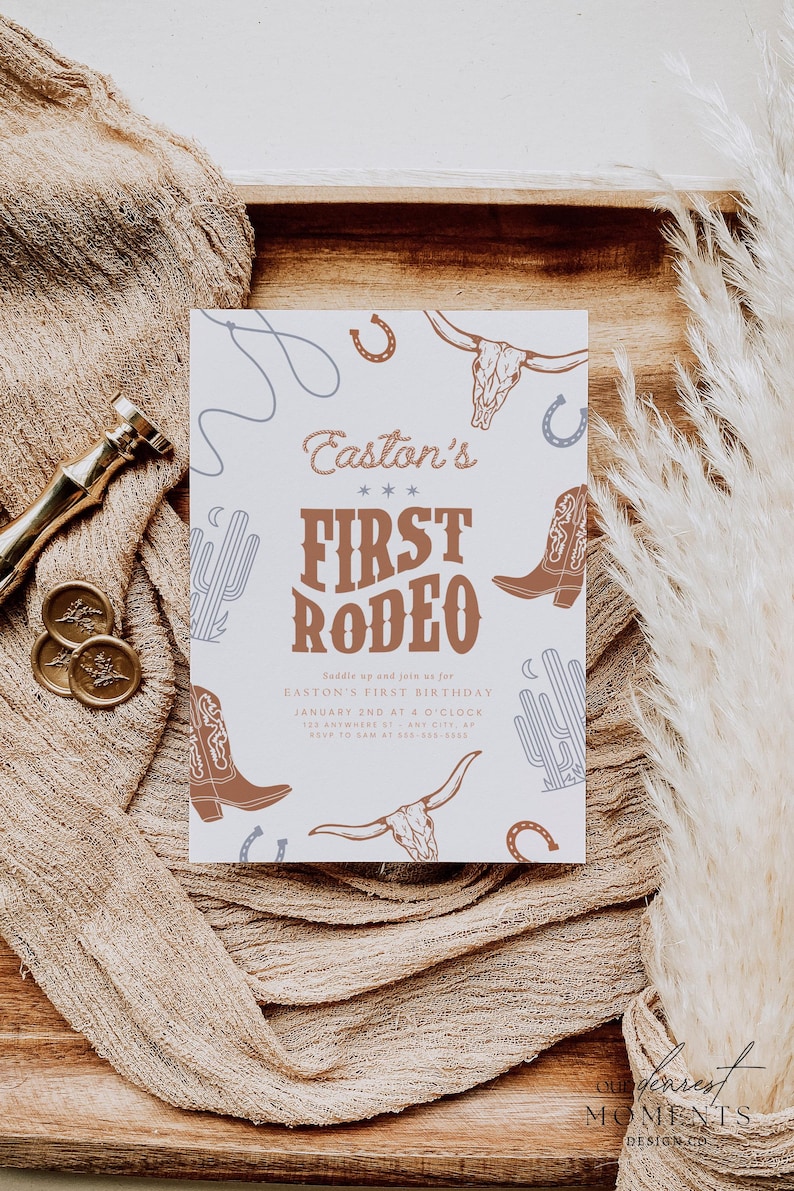 May include: A white invitation card with a rodeo theme. The text "Easton's First Rodeo" is displayed in brown, along with illustrations of cowboy boots, cacti, and horseshoes. The card is on a wooden tray with a beige fabric.