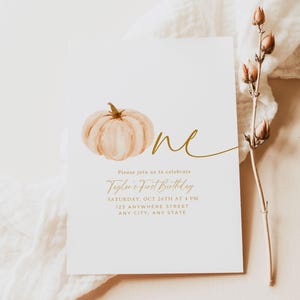 Pu&ograve; includere: Un biglietto d'invito bianco con un'illustrazione di zucca ad acquerello e testo dorato. Il testo recita "One", "Please join us to celebrate Taylor's First Birthday" e i dettagli dell'evento. Il design include il logo "Our Dearest Moments Design Co.".