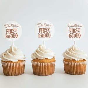 May include: Three white cupcakes with brown frosting and a white circle topper that says "Easton's First Rodeo" in brown text.