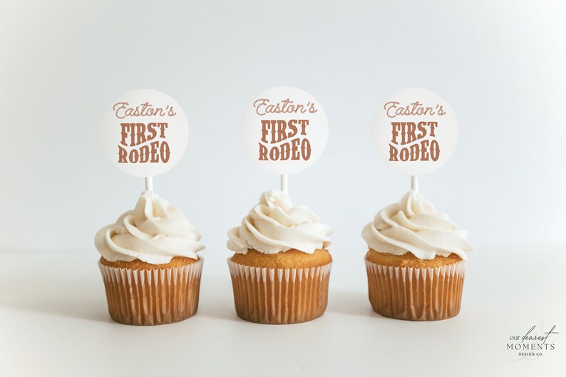 May include: Three white cupcakes with brown frosting and a white circle topper that says "Easton's First Rodeo" in brown text.
