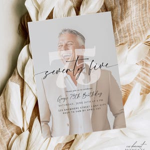 May include: A white and gray invitation card with a photo of a man smiling. The card says "seventy five" in black script and "Greg's 75th Birthday" in black text. The card also includes the date, time, and location of the event.