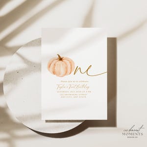 Pu&ograve; includere: Un biglietto d'invito bianco con un'illustrazione di zucca ad acquerello e testo dorato. Il testo recita "One", "Taylor's First Birthday" e i dettagli dell'evento. Il design include le parole "Our Dearest Moments Design Co."