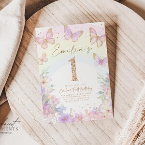 May include: A pastel-colored invitation card for a first birthday celebration. The card features the name "Emilia's" in gold script, a large glittery gold number "1", and delicate butterfly and floral illustrations. The card includes event details.