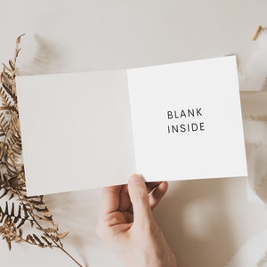 May include: A white card with the text "BLANK INSIDE" written on it. The card is being held open by a hand, with a dried fern branch and white ribbon on a light beige background.