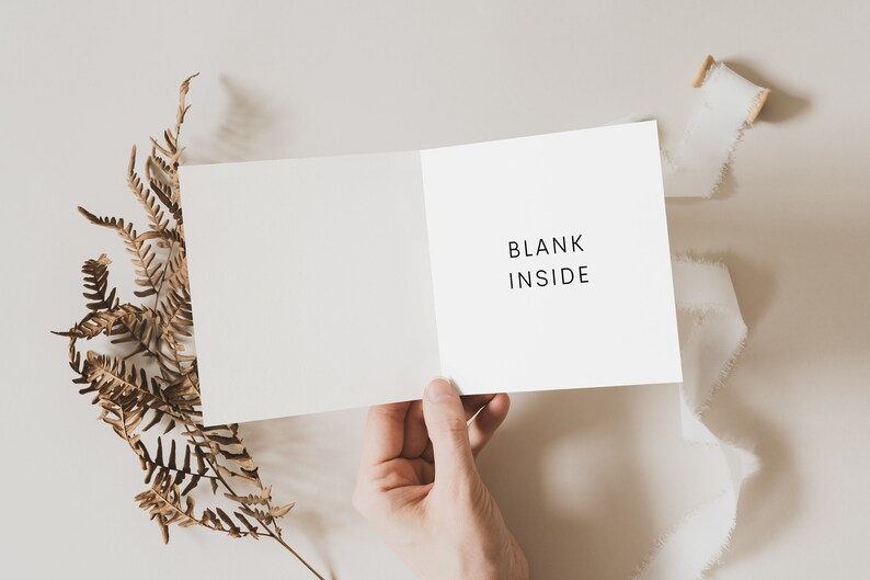 May include: A white card with the text "BLANK INSIDE" written on it. The card is being held open by a hand, with a dried fern branch and white ribbon on a light beige background.