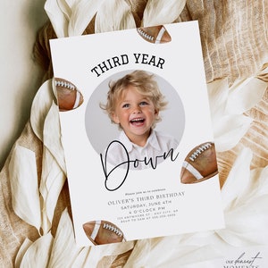 May include: A white invitation with a photo of a young boy in a white shirt. The invitation is decorated with brown and white footballs and the text "THIRD YEAR Down". The text also includes the details of the birthday party: Oliver's Third Birthday, Saturday, June 4th, 4 o'clock PM, 123 Anywhere St, Any City, AP, RSVP to Sam at 555-555-5555. The bottom of the invitation has the text "our dearest MOMENTS DESIGN CO."
