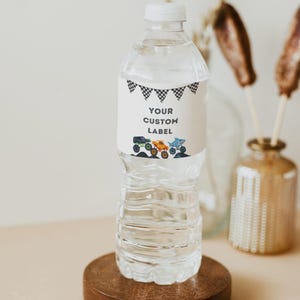 May include: A clear plastic water bottle with a white label featuring the text "YOUR CUSTOM LABEL" and illustrations of monster trucks. The label also has a black and white checkered flag design. The bottle sits on a round wooden base.