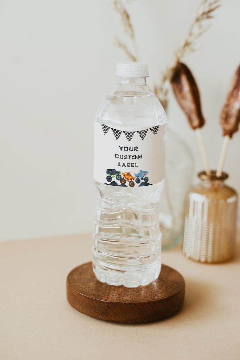 May include: A clear plastic water bottle with a white label featuring the text "YOUR CUSTOM LABEL" and illustrations of monster trucks. The label also has a black and white checkered flag design. The bottle sits on a round wooden base.
