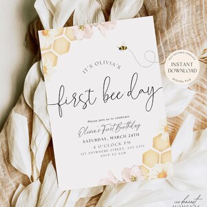 May include: A printable invitation for a first birthday party with a bee theme. The invitation features a watercolour illustration of a honeycomb, flowers, and a bee. The text reads "It's Olivia's First Bee Day" and includes details about the party, such as the date, time, and location. The invitation also includes the text "Our Dearest Moments Design Co."
