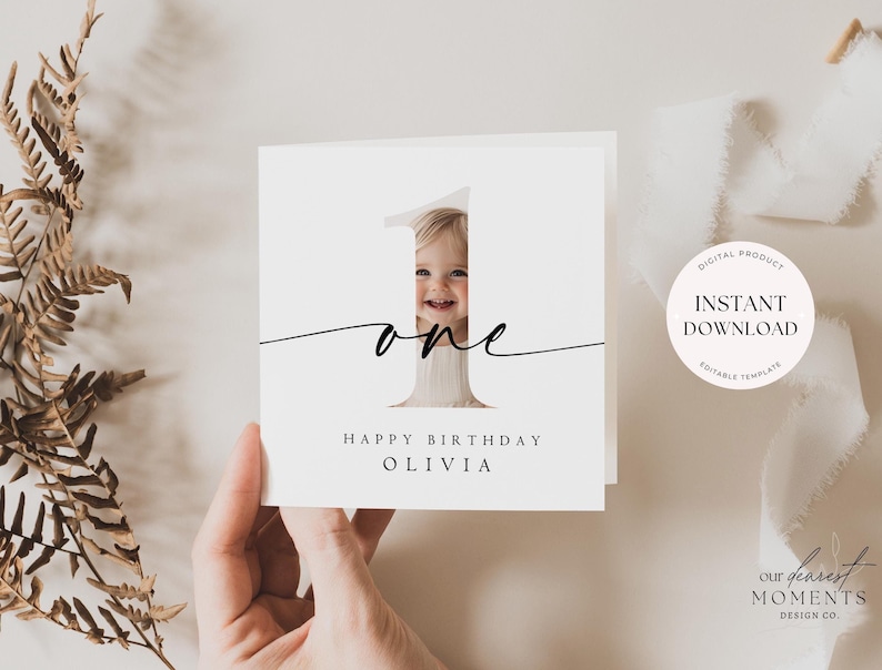 May include: A white card with a black number one with a photo of a baby in the centre. The text "one" is written in cursive below the number. The text "Happy Birthday Olivia" is written below the number.