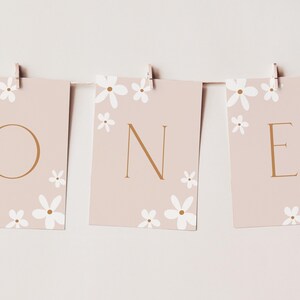 May include: A banner spelling "ONE" for a first birthday celebration. Each card is a light pink colour, adorned with white flower illustrations and gold lettering. The cards are held by wooden clips on a string.