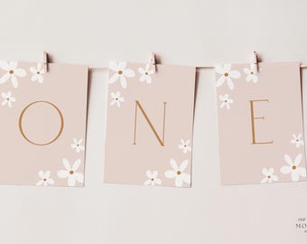 Op de afbeelding: Een slinger met de tekst "ONE" voor een eerste verjaardag. Elke kaart is lichtroze, versierd met witte bloemillustraties en gouden letters. De kaarten worden met houten clips aan een touwtje bevestigd.