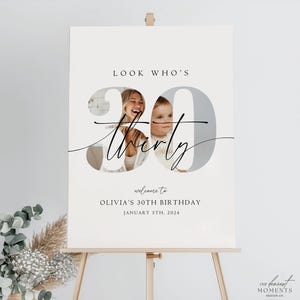 Op de afbeelding: Een wit welkomsbord met een foto van een vrouw en een kind in het getal 30. De tekst luidt "Look Who's Thirty" en "Welcome to Olivia's 30th Birthday January 5th, 2024".