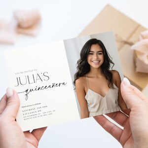 K&ouml;nnte beinhalten: Eine Quincea&ntilde;era-Einladungskarte, die von H&auml;nden gehalten wird, mit einem Foto einer l&auml;chelnden jungen Frau. Der Text der Karte lautet "YOU'RE INVITED TO JULIA'S quincea&ntilde;era" mit Veranstaltungsdetails. Eine Geschenkbox mit Schleife befindet sich im Hintergrund.