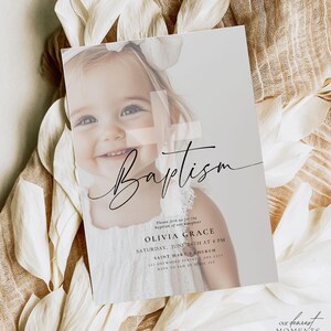May include: A baptism invitation card featuring a smiling child with a white bow. The card has the word "Baptism" in elegant script, along with event details: Olivia Grace, June 26th at 4 PM, and Saint Mary's Church.
