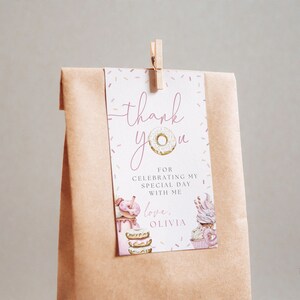 May include: A pink and white thank you tag with a donut illustration and the text "thank you" and "love, Olivia". The tag is attached to a brown paper bag with a wooden clothespin.