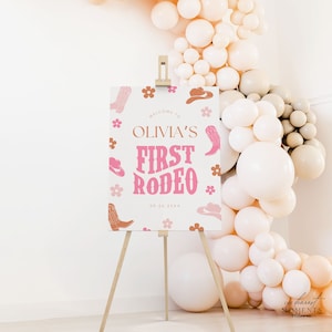 May include: A white sign with pink and brown cowboy boots, hats, and flowers. The sign says "Welcome to Olivia's First Rodeo 09.26.20XX".