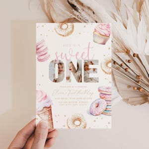 May include: A pink and gold first birthday invitation with a donut and cupcake theme. The invitation features a photo of a child in the number "one" and the text "She's a sweet one".