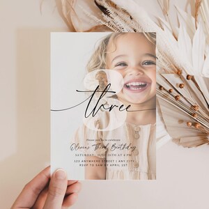 May include: A clear, white invitation card with a photo of a young girl smiling. The card has black calligraphy that says "three" and the text "Olivia's Third Birthday" with details about the event.