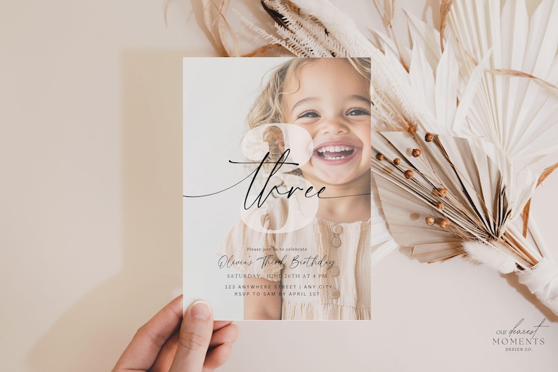 May include: A clear, white invitation card with a photo of a young girl smiling. The card has black calligraphy that says "three" and the text "Olivia's Third Birthday" with details about the event.