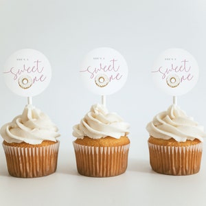 May include: Three cupcakes with white frosting and pink and gold "She's a Sweet One" donut themed cupcake toppers.