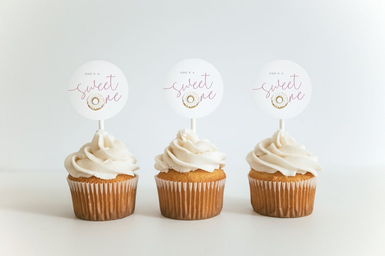 May include: Three cupcakes with white frosting and pink and gold "She's a Sweet One" donut themed cupcake toppers.
