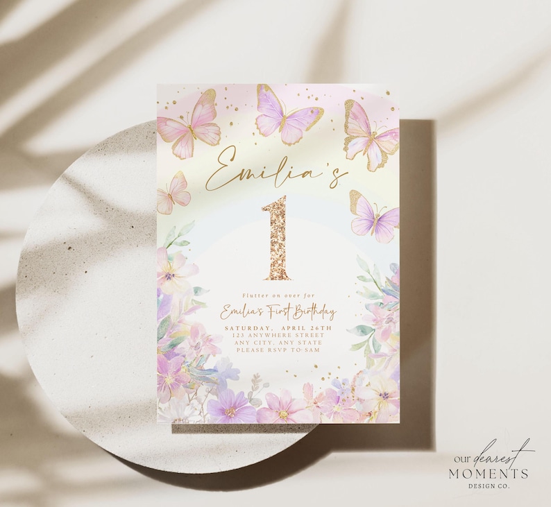 May include: A pastel-colored invitation card with butterflies and floral accents. The card features the name "Emilia's" and the number "1" in gold glitter. Text reads "Emilia's First Birthday" with event details.