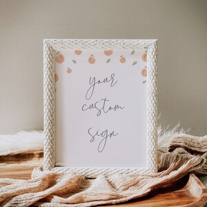 May include: A white frame with a peach and leaf design surrounds a white sign with the text "your custom sign". The frame is sitting on a wooden surface with a tan fabric draped over it.