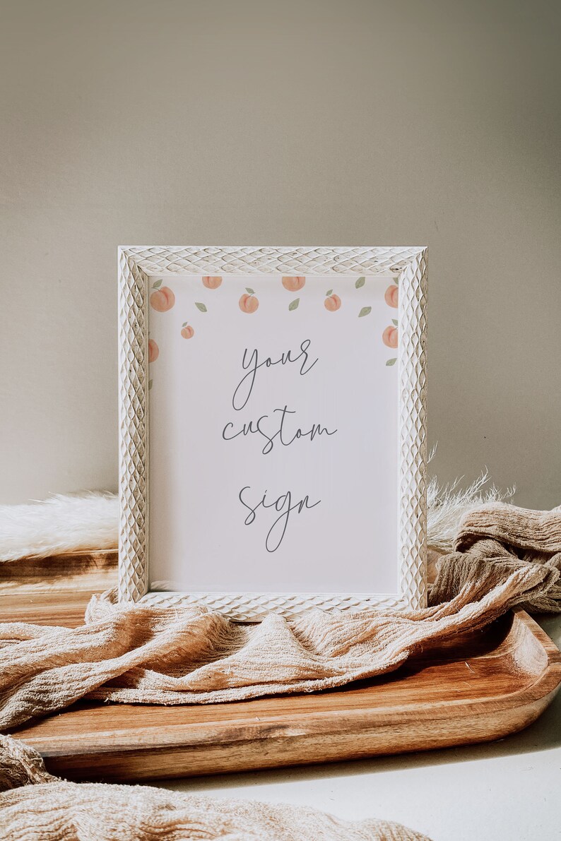 May include: A white frame with a peach and leaf design surrounds a white sign with the text "your custom sign". The frame is sitting on a wooden surface with a tan fabric draped over it.