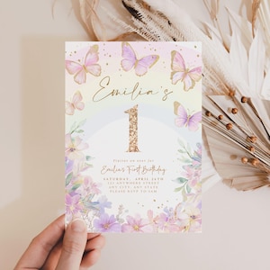 May include: A pastel-colored invitation for a first birthday, featuring butterflies and floral accents. The invitation reads "Emilia's 1st Birthday" with the date and location details. The number "1" is embellished with gold glitter.