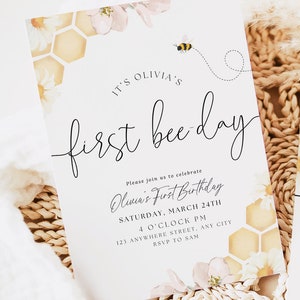 May include: A white invitation card featuring a bee and honeycomb design. The text on the card reads "It's Olivia's First Bee Day." The invitation details a first birthday party for Olivia, including the date, time, and location.