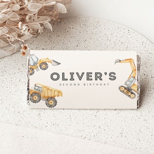 May include: A white card with the text "Oliver's Second Birthday" in black. The card is decorated with yellow construction vehicles, including a dump truck, an excavator, and a bulldozer.