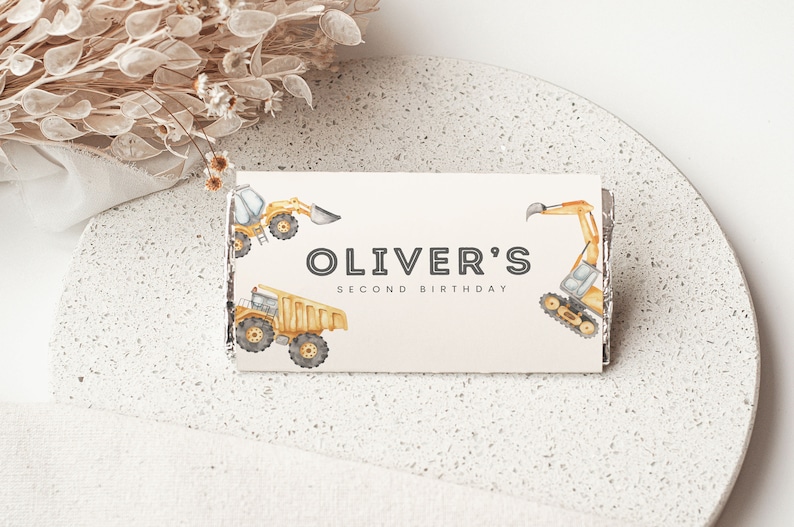 May include: A white card with the text "Oliver's Second Birthday" in black. The card is decorated with yellow construction vehicles, including a dump truck, an excavator, and a bulldozer.