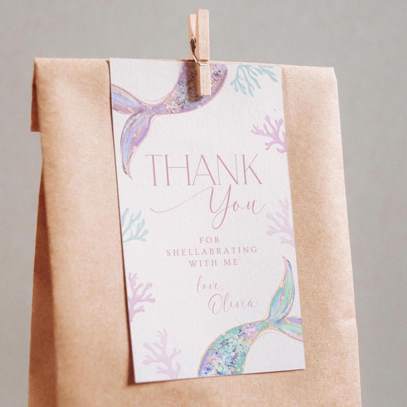 May include: A thank you tag with a mermaid tail design. The tag is white with pink and blue accents. The text on the tag reads "Thank You for celebrating with me, love, Olivia".