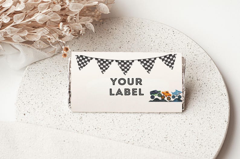 May include: A rectangular label featuring the words "YOUR LABEL" in black, decorated with a checkered flag garland and illustrations of monster trucks and a shark. The label is on a white, speckled surface.