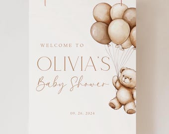 Könnte beinhalten: Ein weißes Willkommens-Schild mit braunem Text, der "Willkommen zu Olivia's Baby Shower 09.26.2024" lautet. Ein brauner Teddybär hält braune Ballons fest.