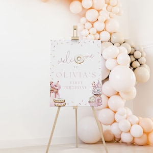 May include: Welcome to Olivia's First Birthday sign with a donut graphic and sprinkles. The sign is on a wooden easel in front of a wall decorated with a balloon arch.