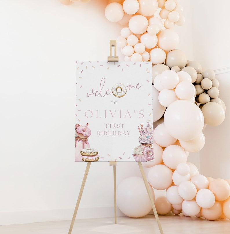 May include: Welcome to Olivia's First Birthday sign with a donut graphic and sprinkles. The sign is on a wooden easel in front of a wall decorated with a balloon arch.