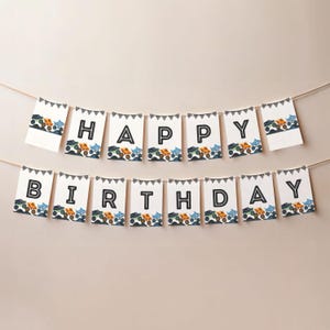 May include: A birthday banner spelling out "HAPPY BIRTHDAY" in black capital letters on white flags. Each flag has a black and white triangle border and a row of colourful monster truck illustrations along the bottom.