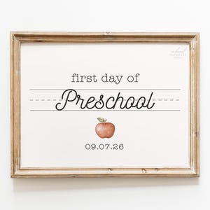 May include: A rectangular sign in a light wood frame featuring the text first day of Preschool in black lettering on a white background. Below the text is a watercolour illustration of a red apple and the date 09.07.26 in black font.