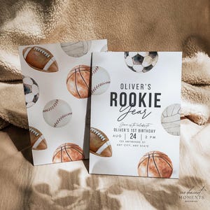 May include: A white invitation with a watercolor illustration of sports balls, including a basketball, baseball, football, soccer ball, and volleyball. The text on the invitation reads "Oliver's Rookie Year" and includes details for a first birthday party.