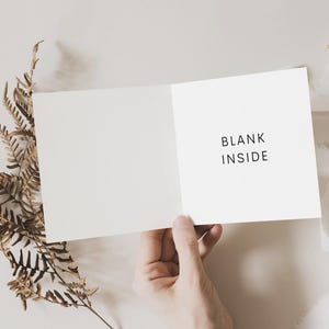 May include: A blank white card with the text "BLANK INSIDE" printed on the front. The card is held open by a hand, and a brown fern is in the background.