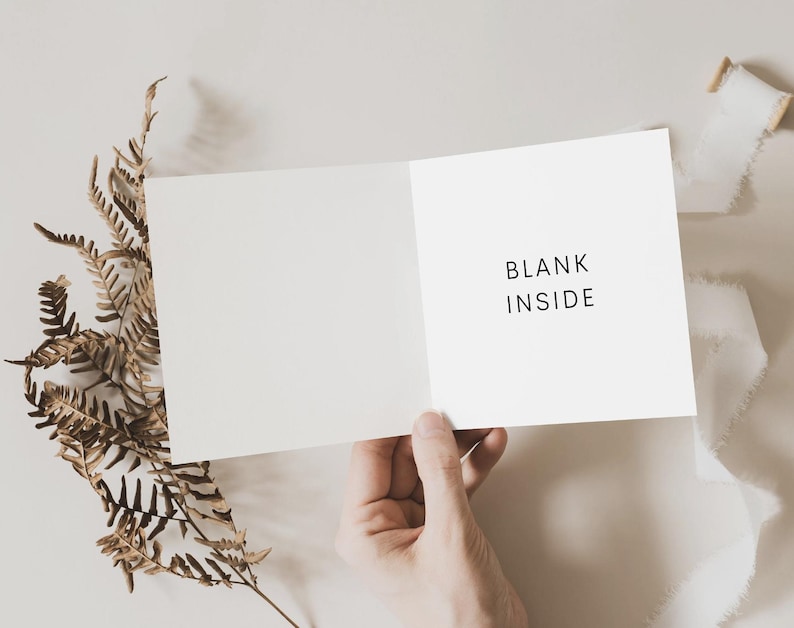 May include: A blank white card with the text "BLANK INSIDE" printed on the front. The card is held open by a hand, and a brown fern is in the background.
