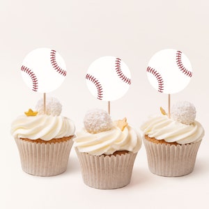 May include: Three white cupcakes with white frosting and gold stars. Each cupcake has a white baseball-shaped topper with red stitching.