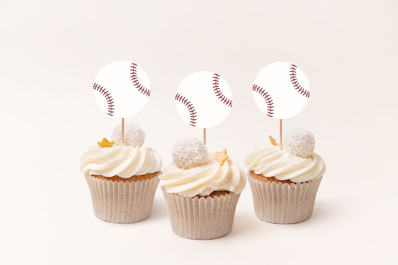 May include: Three white cupcakes with white frosting and gold stars. Each cupcake has a white baseball-shaped topper with red stitching.