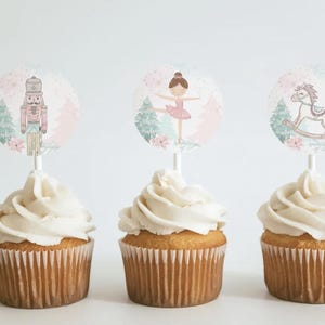 May include: Three cupcakes with white frosting and decorative toppers. The toppers feature illustrations of a nutcracker, a ballerina, and a rocking horse. The cupcakes are in brown paper liners, perfect for a themed party or event.