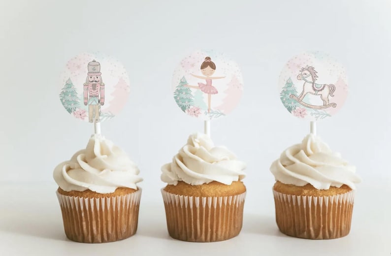 May include: Three cupcakes with white frosting and decorative toppers. The toppers feature illustrations of a nutcracker, a ballerina, and a rocking horse. The cupcakes are in brown paper liners, perfect for a themed party or event.