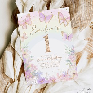 May include: A pastel-colored invitation with butterflies and floral accents. The invitation reads "Emilia's First Birthday" with the number "1" in gold glitter. The design includes the event details and the text "Our Dearest Moments Design Co."