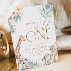 May include: A white invitation card with a gold and white "Wild One" design. The card features a giraffe, elephant, and koala with floral crowns. The text reads "Olivia's a Wild One" and "Please join us to celebrate Luna's First Birthday, Saturday, June 26th at 4 PM, 123 Anywhere Street, Any City, Any State, RSVP to Sarah". The bottom of the card reads "Our Dearest Moments Design Co."