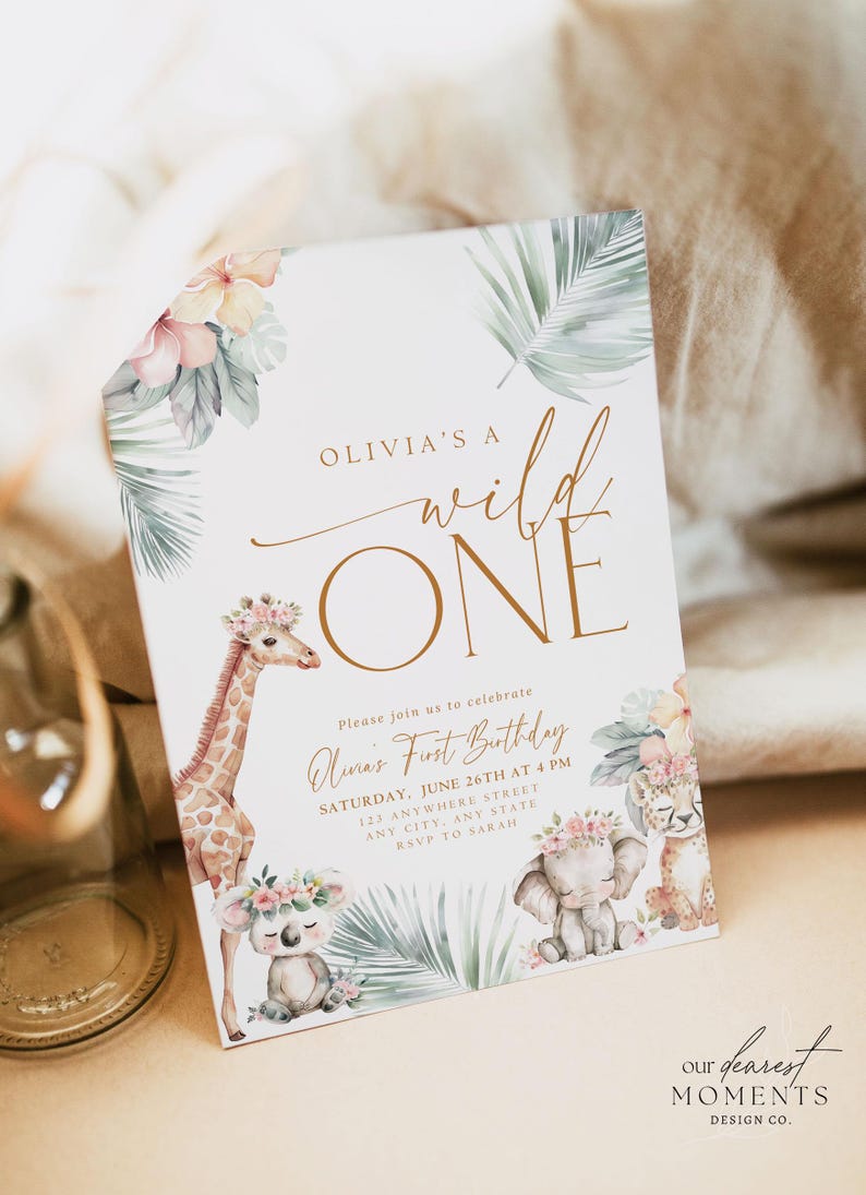 May include: A white invitation card with a gold and white "Wild One" design. The card features a giraffe, elephant, and koala with floral crowns. The text reads "Olivia's a Wild One" and "Please join us to celebrate Luna's First Birthday, Saturday, June 26th at 4 PM, 123 Anywhere Street, Any City, Any State, RSVP to Sarah". The bottom of the card reads "Our Dearest Moments Design Co."